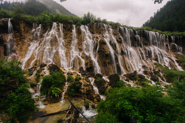 A dramatic waterfall cascades down a steep, moss-covered cliff face in a misty, mountainous landscape. © JX