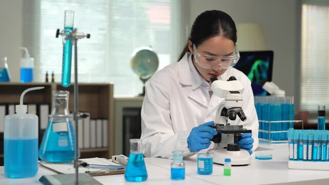 Young female asian scientist in a lab coat and goggles adjusting a microscope slide while conducting a medical experiment for pharmaceutical research and development