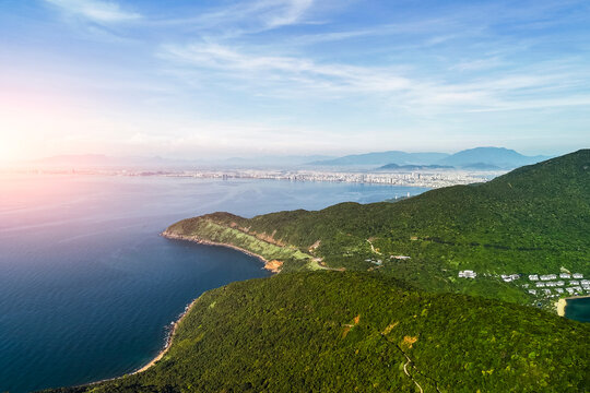Dense green jungle foliage blankets mountain terrain extending to a peaceful blue ocean. A coastal city lines the horizon under a bright daytime sky
