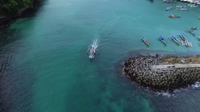 Aerial drone footage of a traditional fishing boat leaving the harbor pier and heading toward the open sea. Cinematic aerial perspective capturing the vessel departing from the dock