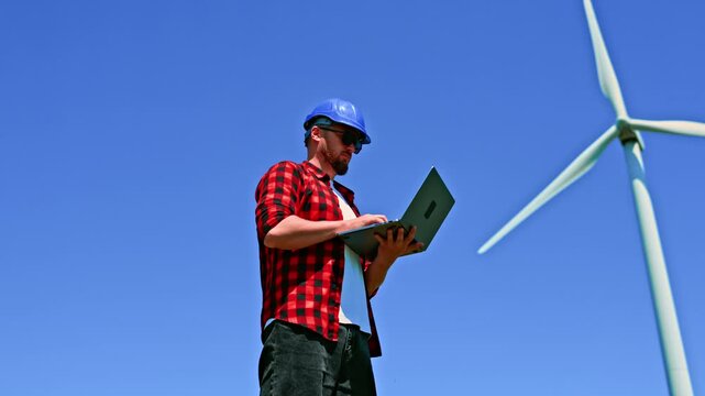 A windmill engineer stands with a laptop near a wind turbine. The sky is blue, and the engineer focuses on their work while wearing a hard hat and sunglasses.