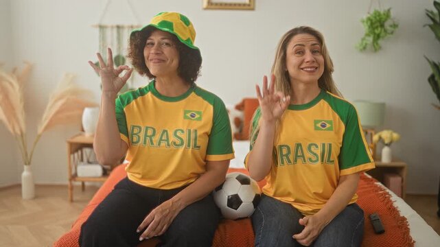 Woman friends seated on bed wearing brasil shirts holding soccer ball and making ok hand sign in building; team pride.
