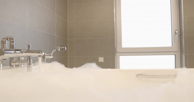 A white bathtub filled with thick white foam in a bathroom with a window and grey tiled walls