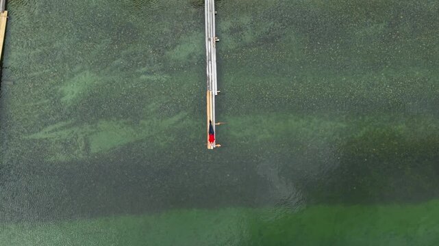 Top down drone aerial view of a male tourist in a red t-shirt lying on a wooden pier and taking a selfie as the camera ascends straight up.