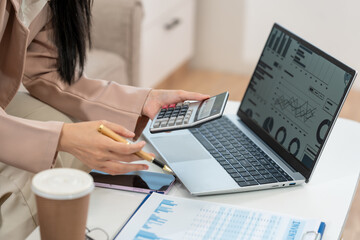 Businesswoman reviewing charts on a laptop while using a calculator and phone at a desk with...
