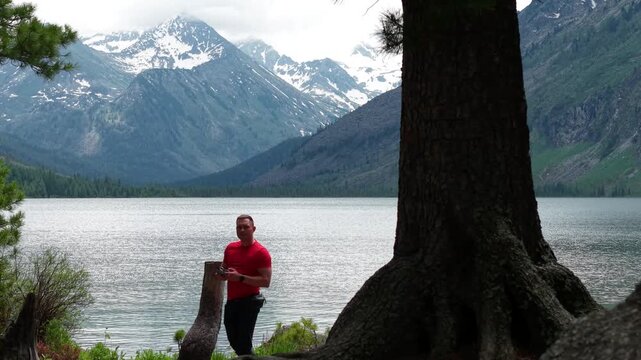 Cinematic drone aerial sideways panning shot passing behind a large cedar tree trunk, creating a natural wipe transition reveal of a mountain lake and snow-capped peaks.