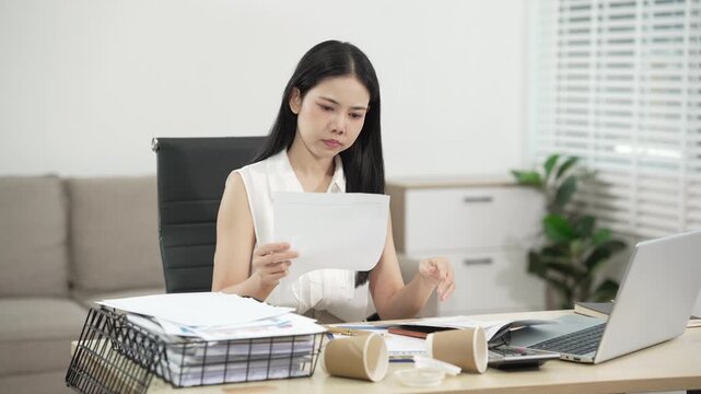 Young woman sorting papers at a desk with laptop, scattered documents and coffee cups in a bright home office, showing stress, overtime, paperwork and busy remote work.