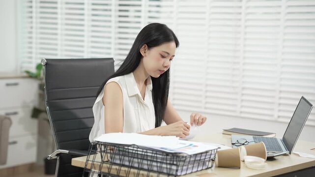 Young woman sorting papers at a desk with laptop, scattered documents and coffee cups in a bright home office, showing stress, overtime, paperwork and busy remote work.
