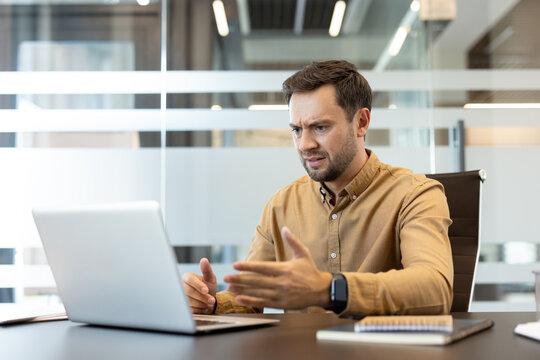 Young businessman experiencing technical difficulties on his laptop, expressing confusion and stress while gesturing at the screen in a contemporary glass office environment