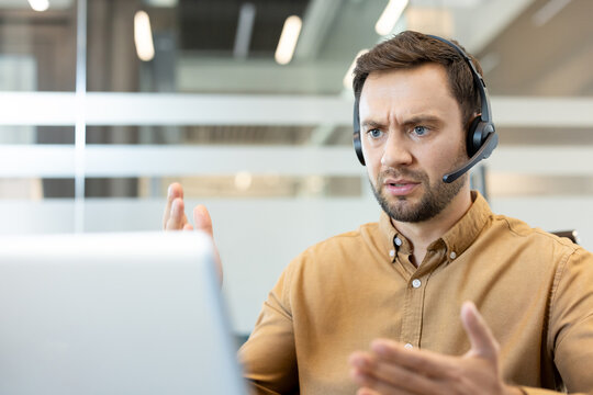 Male customer support agent with a headset responding to a challenging call, showing feelings of anger and stress while working on a laptop in a modern office setup