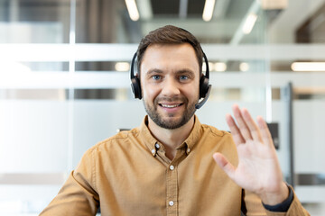 Obraz premium Man wearing a headset with a microphone, smiling confidently and waving hand while communicating during a remote business video call from an office