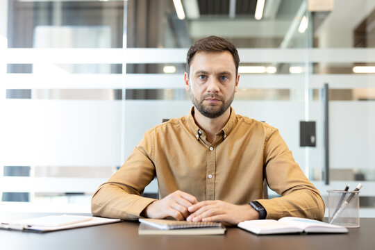 Professional man with a serious expression sitting at his desk in a bright, modern office, facing forward directly into the camera during a video conference or virtual meeting