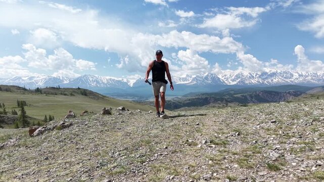 Low altitude drone shot of an athletic man confidently walking forward while holding a remote controller against a scenic snowy mountain range background.