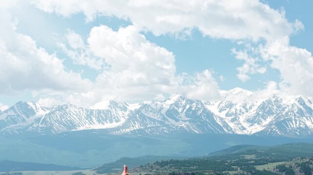 An athletic male adventurer sitting cross-legged on a mountain peak and pointing at a majestic snow-capped mountain range before giving a thumbs up gesture.