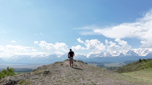 Young male traveler from behind walking down a green hill trail toward a majestic panoramic mountain range under a blue cloudy sky