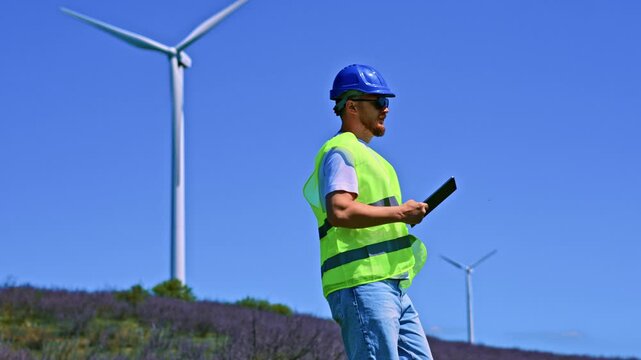 A windmill engineer stands in a green safety vest looking at a wind turbine. The engineer holds a tablet and checks the equipment in an open field during daytime.