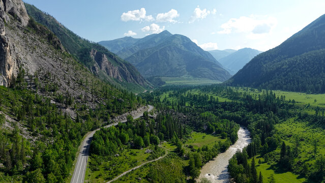High-angle drone aerial view of the Chuysky Tract highway winding through a lush mountain river valley near Aktash in Altai.