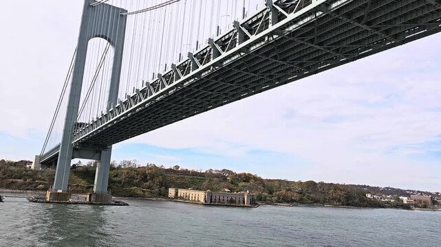 Horizontal low angle shot of the massive Verrazzano Narrows suspension bridge spans over water in New York, with fort Wadsworth and autumn trees in the background. High quality FullHD footage