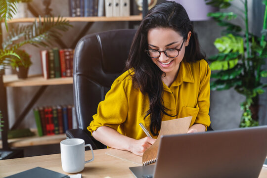 Young smiling woman freelancer sitting and writing in planner indoors in cozy home office, female entrepreneur planning tasks, remote work productivity concept