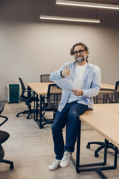 Smiling middle aged man wearing eyeglasses with beard showing thumbs up in modern office