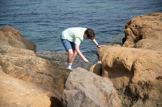 Teenage boy exploring the rocky shoreline of Tabarca while searching among coastal rocks on a sunny summer morning by the Mediterranean Sea