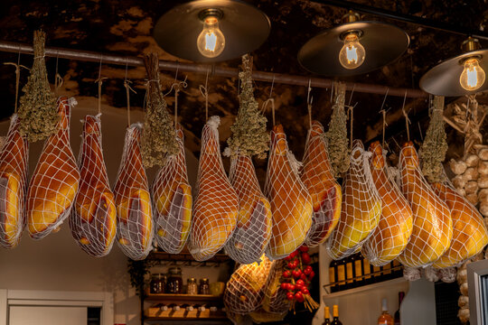 Traditional Italian cured hams hanging in a delicatessen with dried herbs and warm lighting. Authentic gourmet food display showcasing artisanal cuisine and Mediterranean flavors