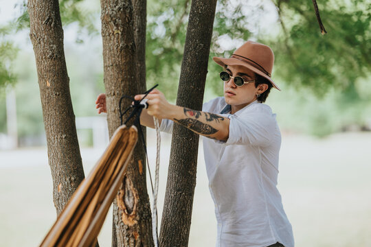 Young man wearing a hat and sunglasses ties a hammock to a tree. He prepares the hammock outdoors among trees in a quiet park setting.
