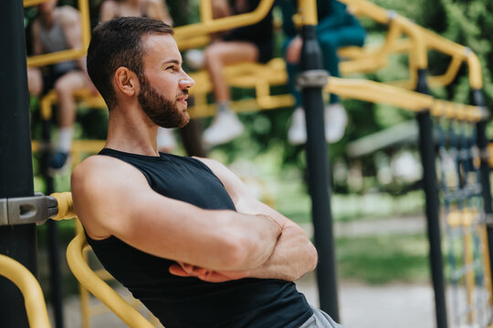 Young man with a beard rests on yellow bars at an outdoor gym. He leans back with arms crossed, taking a break during a park workout.