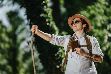 Young male hiker wearing a hat and sunglasses holds a notebook and walking stick in a sunlit forest. The traveler pauses with a backpack, looking upward while standing among trees.