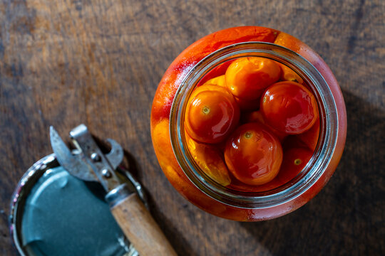 Canned red pickled cherry tomatoes inside an open glass jar on rustic wooden table with an old can opener, top view, closeup. Flat lay food photography for grocery store promos or cooking recipes