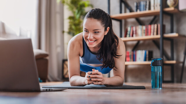 Young woman training at home, doing plank exercise and watching videos on laptop, fit female doing online workout, fitness, wellness and healthy lifestyle concept