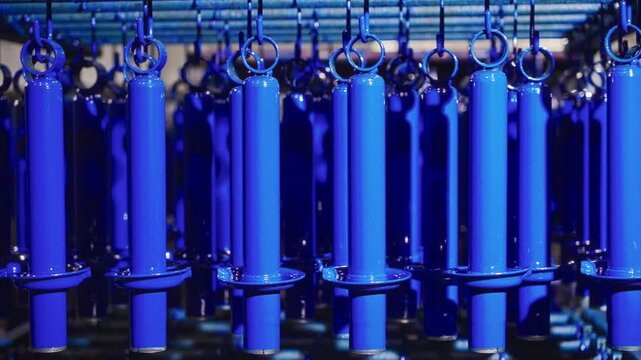 Rows of cylindrical metal components hanging from overhead hooks receiving fresh vivid blue powder coating on factory conveyor line with glossy finish and bright specular highlights