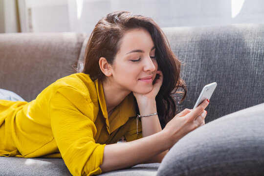Relaxed smiling woman on couch using smartphone and texting at home, happy female chatting online, social media, communication and cozy lifestyle concept