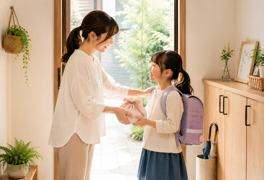 母親が手作りのお弁当を小学生の娘に渡す朝の玄関風景