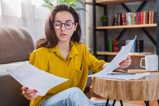 Young mixed race woman in eyeglasses holding paper bill letter at home, female reading invoice, managing household expenses and financial planning concept