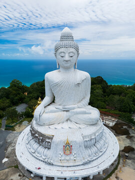 Aerial view of the white marble Big Buddha statue seated on a hilltop, surrounded by lush green trees with the blue Andaman Sea in the background in Phuket, Thailand.