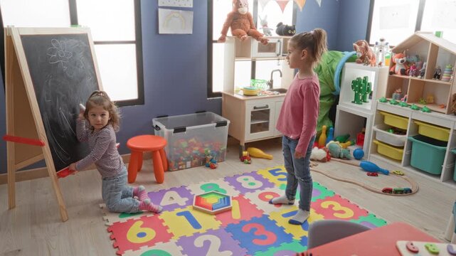 Two girls, one kneeling with chalk at a freestanding chalkboard while the other stands with foot on a wooden rainbow stacker in a playroom building; playful curiosity.