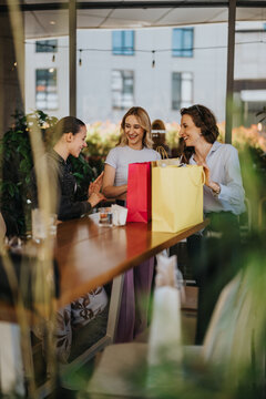 Three female friends celebrate with shopping bags at a cafe table. They smile and chat while holding colorful gift bags.