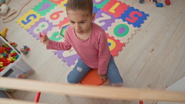 Girl kneeling and drawing with pink chalk on a small easel near a foam number playmat and box in building; playful learning.