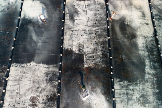 Three people manually collect white salt from expansive, dark salt pans, forming distinct white piles. Shadows stretch across the cracked ground