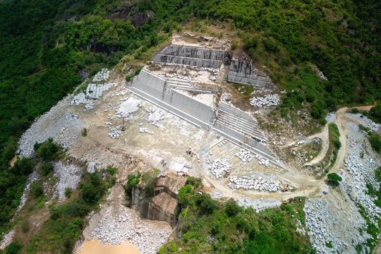 Extensive rock quarry operates on a verdant mountain, displaying stepped retaining walls, piles of cut stone blocks, and muddy water below