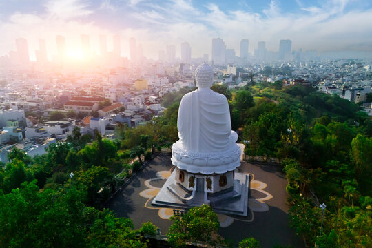 White Buddha atop Long Son Pagoda hill watches Nha Trang city awaken at sunrise, warm light painting the urban scene and lush trees
