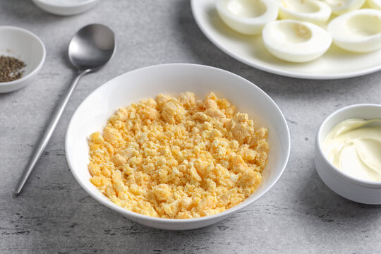 Crumbled hard boiled egg yolks in a bowl, garlic, mayonnaise and seasonings, prepared as filling for stuffed eggs on a gray textured surface