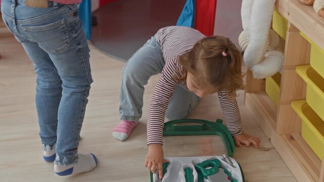 Girl kneels and reaches hand into green plastic toy toolkit on pale wood floor beside yellow storage shelving inside a building; curiosity play exploration.
