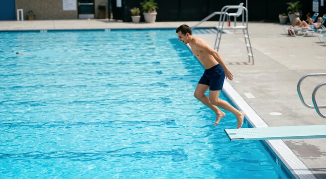 Man jumping off a diving board into a blue swimming pool. Male swimmer in mid-air enjoying summer water activities at an outdoor pool facility. Active vacation lifestyle