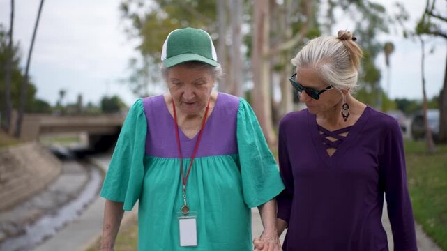 Daughter visits her mother at the care facility. They walk in the park sharing smiles and stories. They enjoy their time together.