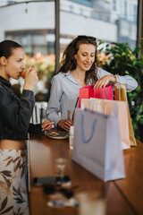 Two women sit at a cafe table with colorful shopping bags and drinks, smiling as they look through purchases. The friends enjoy a casual shopping break and share a joyful moment together.