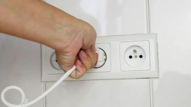 Close-up of a female hand unplugging an electrical power plug from a wall socket. Everyday household action illustrating electrical safety, energy saving, disconnecting electronic devices.