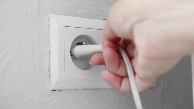 Close-up of a female hand plugging an electrical power plug into a wall socket. Everyday household action showing connecting an electronic device to the power supply, home electricity.