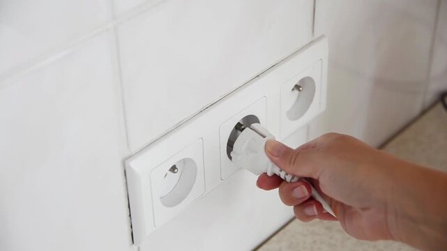 Close-up of a female hand plugging an electrical power plug into a wall socket. Everyday household action showing connecting an electronic device to the power supply, home electricity.
